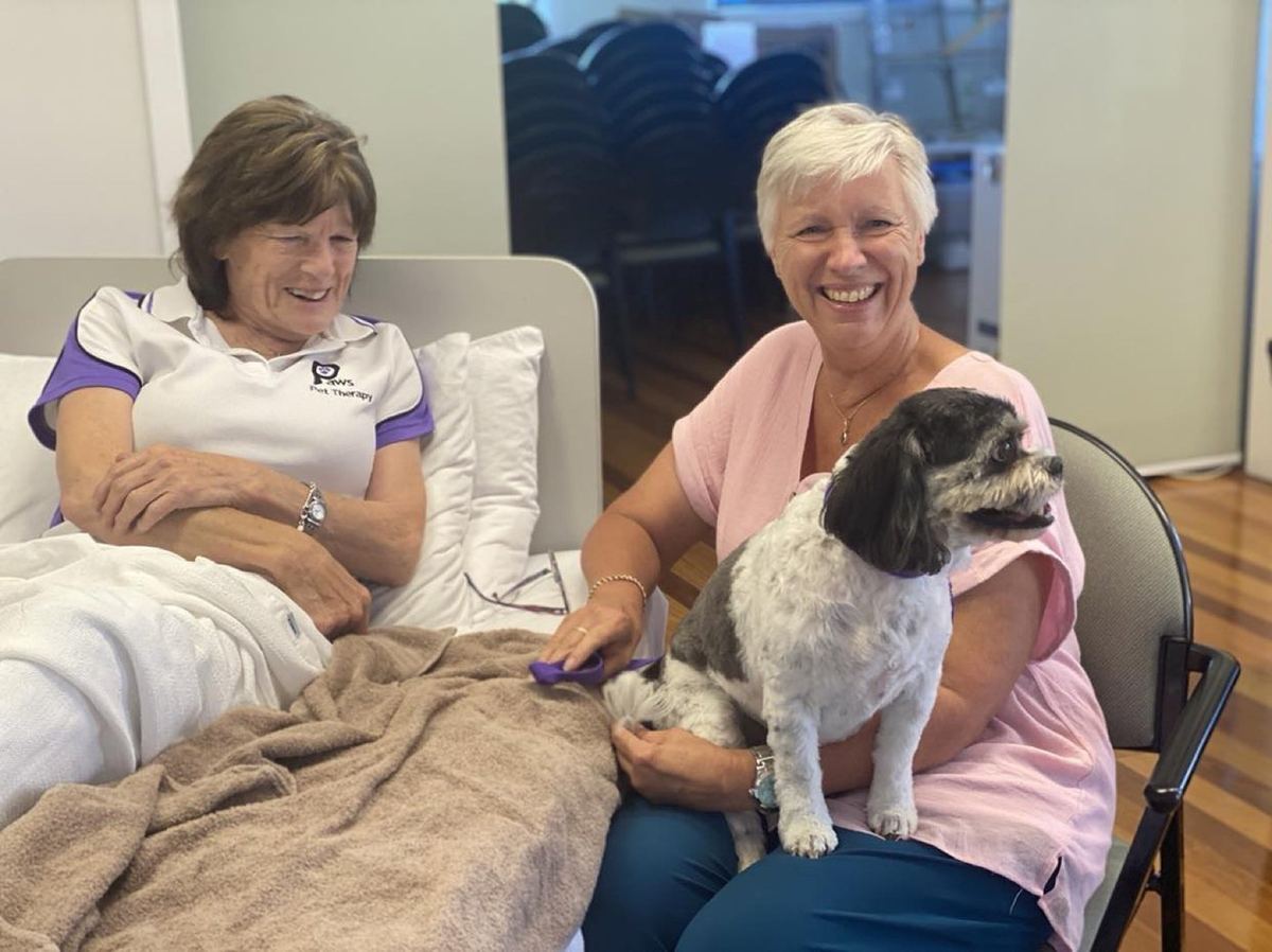 A PAWS volunteer and her small fluffy therapy dog visiting a smiling resident in aged care